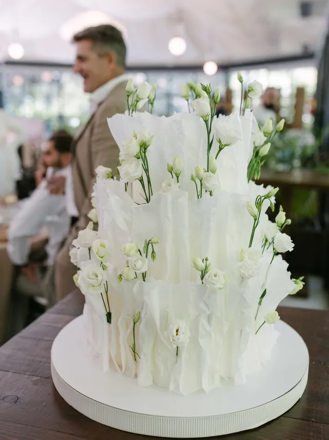 La tarta de lisianthus en su mesa, con invitados de la boda al fondo de un invernadero.