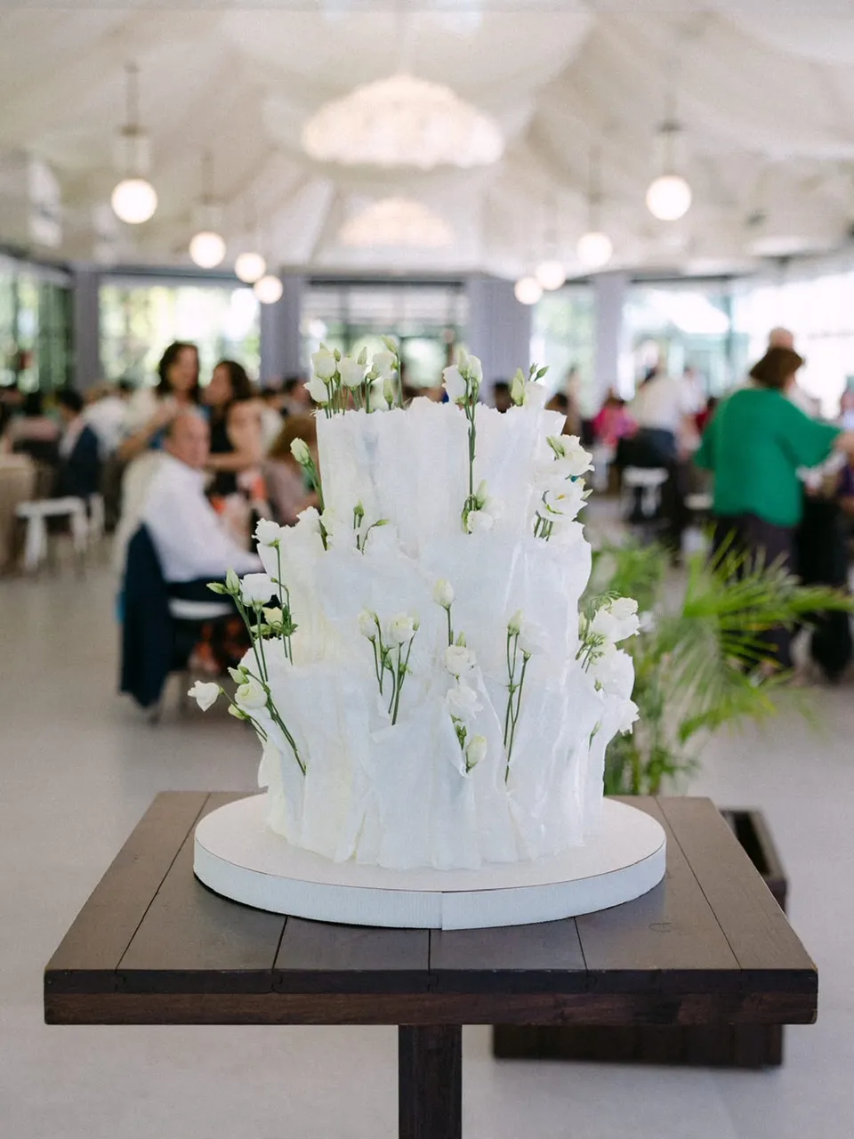 Vista amplia de la tarta de lisianthus sobre una mesa oscura dentro del invernadero, con el lugar de la celebración alrededor.