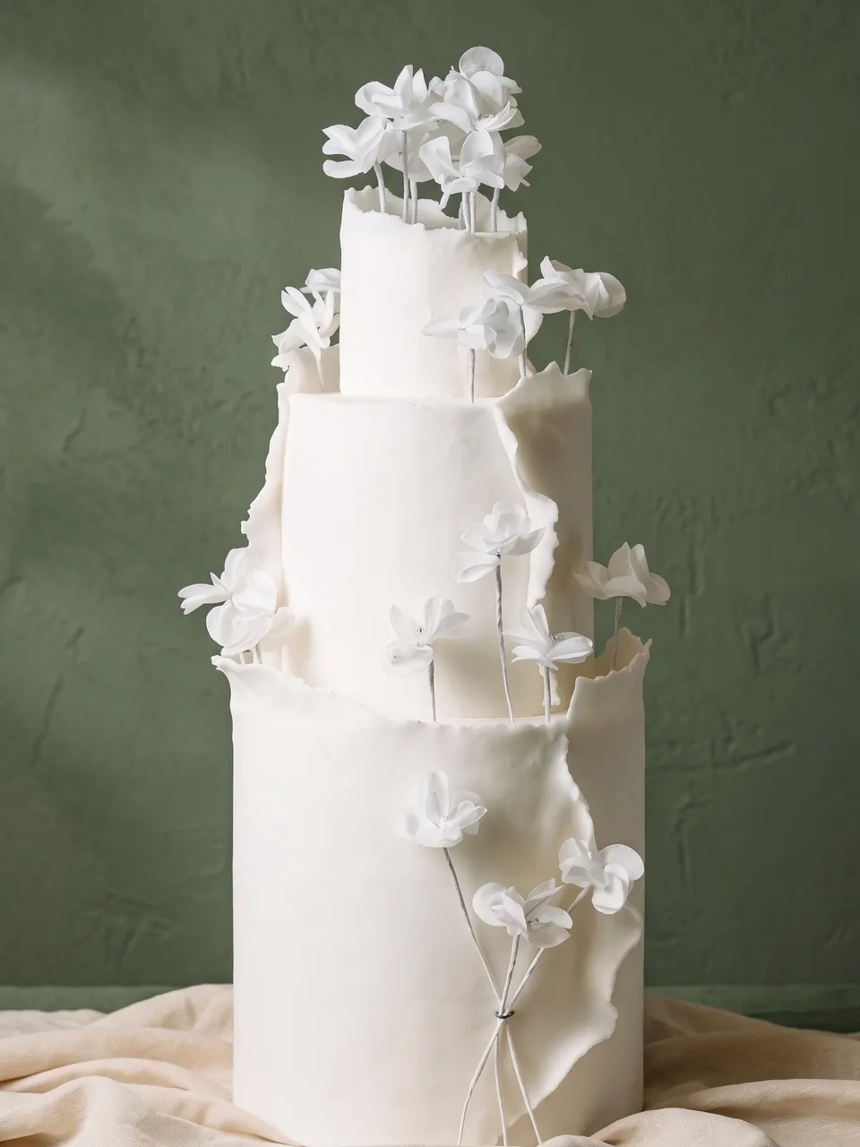 Tarta blanca de tres pisos con bordes rasgados y flores de azúcar blancas con alambre, con los tallos atados en la base, sobre un paño de lino.