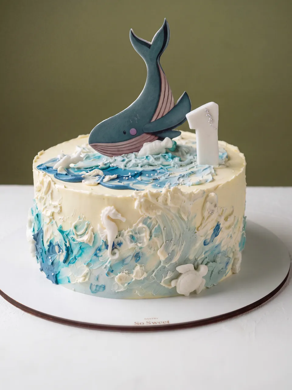 Tarta de primer cumpleaños con una ballena y un número uno encima, y olas de crema de mantequilla azul y blanca con un caballito de mar y una tortuga en el lateral.