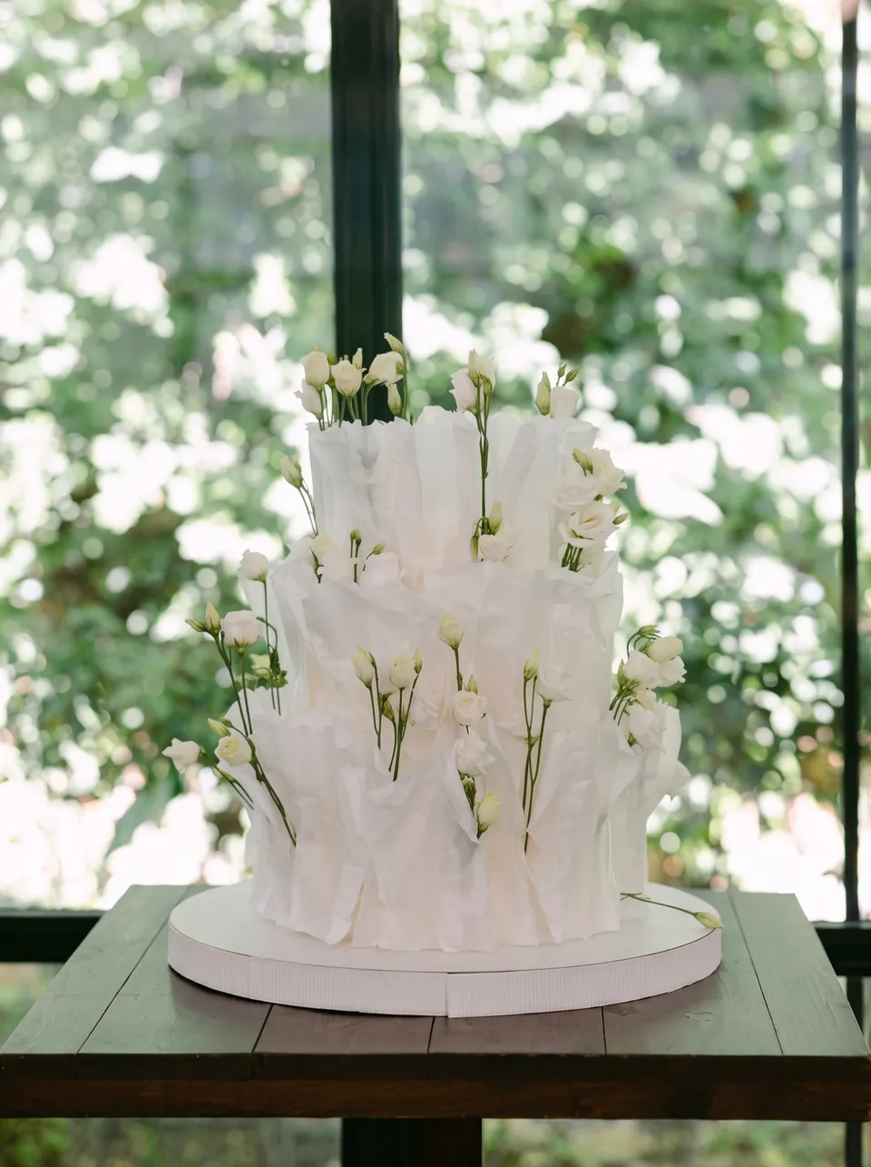 Tarta blanca texturizada de tres pisos con lisianthus blancos frescos, sobre una mesa oscura frente a una ventana de invernadero.