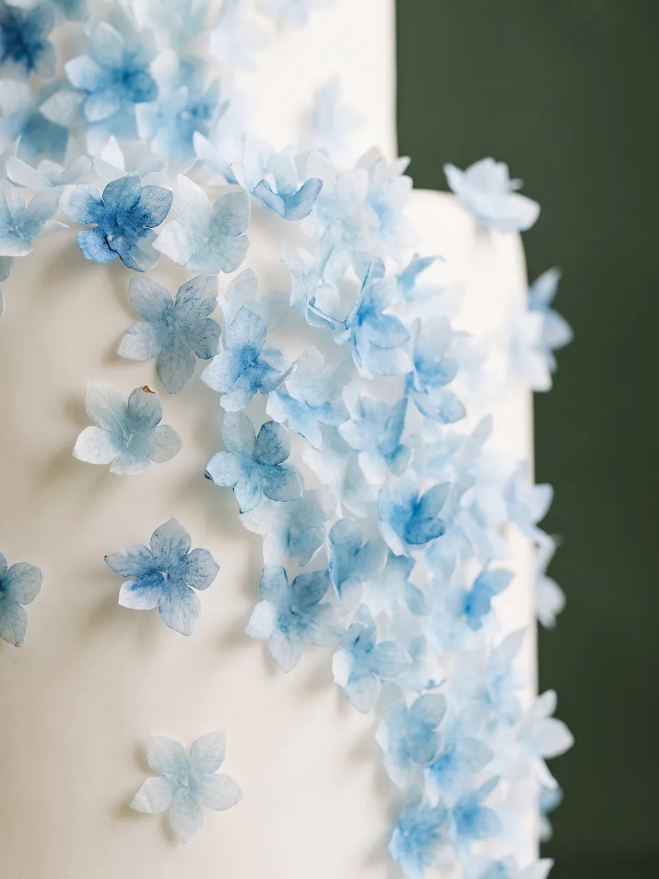 Tarta nupcial blanca de dos pisos con una cascada de flores de azúcar azul pálido, sobre un soporte color crema frente a una pared verde oscuro.